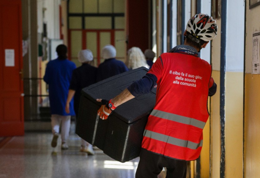 A man wearing a red vest and a bicycle helmet carries a food container in his hands.